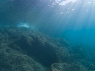 Dramatic underwater landscape off the French coast, where bright sunbeams cut through the deep blue water to illuminate a dark, rocky seabed.