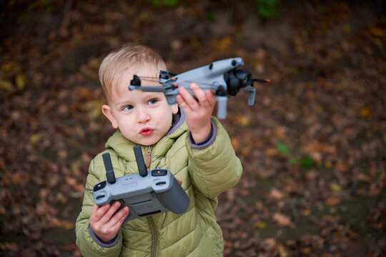 A curious young boy holds a drone in one hand and a remote controller in the other, exploring nature among colorful autumn leaves. His excitement fills the air.