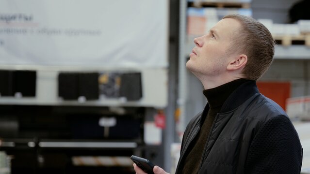 Warehouse supervisor is gazing at the ceiling while holding a smartphone, deep in thought about the day's responsibilities. A man in a hardware store, choosing flooring, shelves with ceramic tiles.
