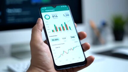 A person analyzing financial data on a smartphone in a modern workspace with charts displayed - Powered by Adobe