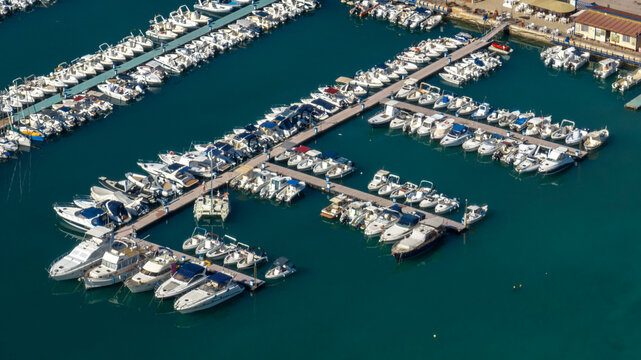 Aerial view of many boats anchored at a pier in a small port. The blue sea surrounds the pier. - Powered by Adobe