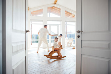 Child on wooden rocking horse in bright room, while adults play and dance, creating joyful atmosphere of family bonding and happiness in spacious interior