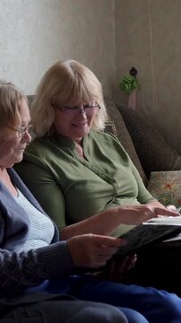 Elder mother and adult daughter looking family vintage photo album sitting on sofa at home. Females sharing cherished family memories. Spending time with family. Visiting elderly in nursing home.