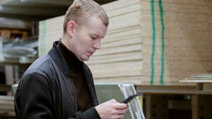 Man using phone to choose construction materials in hardware store for diy project. Man in a hardware store, choosing lumber, shelves with lumber
