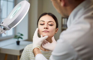 Unrecognizable dermatologist examining patient face, directing lamp for good vision, posing in...