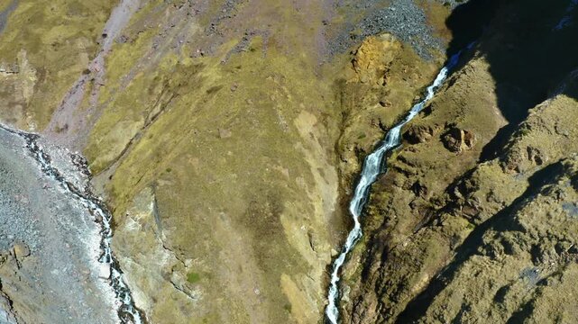 An aerial drone shot captures the stunning natural landscape of the North Caucasus mountains in Russia. A clear stream cascades down a rugged, grassy slope, highlighting the untouched beauty of the re