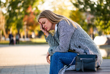Portrait of exhausted businesswoman in coat having a headache while sitting alone on bench in the city park.