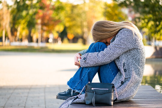 Portrait of sad and despair businesswoman in coat sitting alone on a bench in the city park during autumn day. - Powered by Adobe