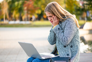 Portrait of stressed and anxious businesswoman in coat using laptop while sitting on a park bench in the city.