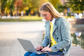Portrait of confident businesswoman with eyeglasses  in coat using laptop while sitting on a park bench in the city during sunny autumn day.