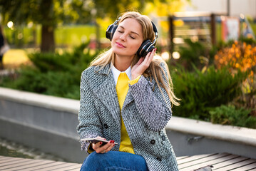 Portrait of beautiful modern businesswoman in autumn coat listening to music through wireless headphones and using smartphone while relaxing on a park bench in the city.