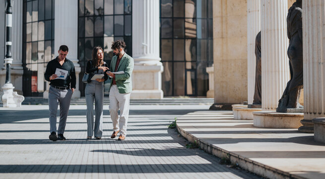 Three people stand outside a stately building, reviewing papers and screens, sharing ideas and plans. A bright, collaborative moment in a modern urban setting with classic architecture.