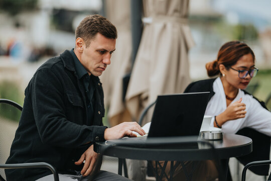 A man concentrates on a laptop while a woman nearby chats, both seated at a round table on an outdoor cafe. Ideal for business, collaboration, or remote work themes. - Powered by Adobe
