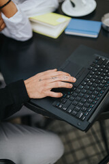 A close-up of a tattooed hand typing on a black laptop at a desk. Colorful notebooks and a coffee cup sit nearby, creating a casual workspace atmosphere.