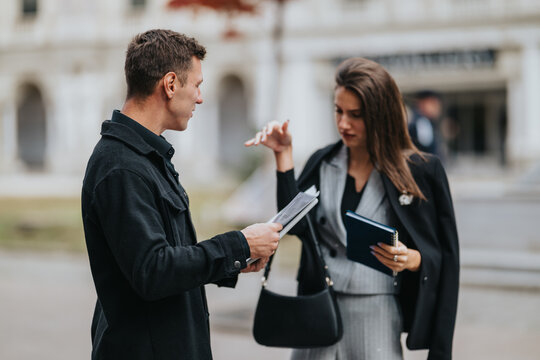 A man and a woman in smart attire converse while reviewing papers and a notebook outside a modern building. The scene captures teamwork, planning, and professional collaboration in an urban area.