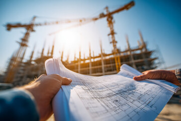Architect holding blueprints at a construction site with cranes and building framework visible under bright sunlight during daytime project planning