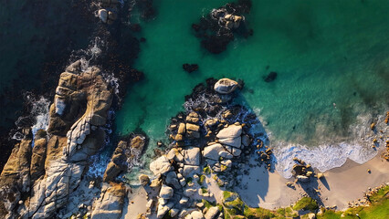 Aerial view of rugged, sun-kissed boulders meeting the turquoise ocean, while the shoreline whispers tales of time, Cape Town, Western Cape, South Africa.