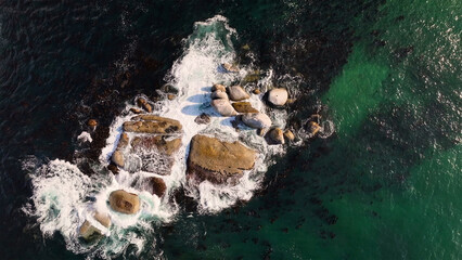 Aerial view of waves crashing against weathered boulders, where the deep blues meet the vibrant greens of the ocean, creating a mesmerizing contrast, Cape Town, Western Cape, South Africa.