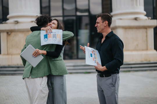 Two coworkers share a warm hug while one holds a tablet and another a chart, as a smiling man stands nearby with a report outside a corporate building, highlighting teamwork and collaboration.