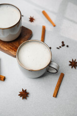 Two warm mugs with milk and cinnamon sticks on a light gray surface with star anise.