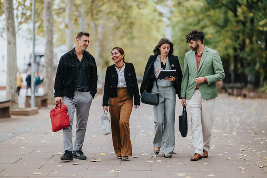 A diverse group of four people walks and talks in a leafy park. They wear smart casual outfits, carry bags and a tablet, and share a friendly, collaborative moment.