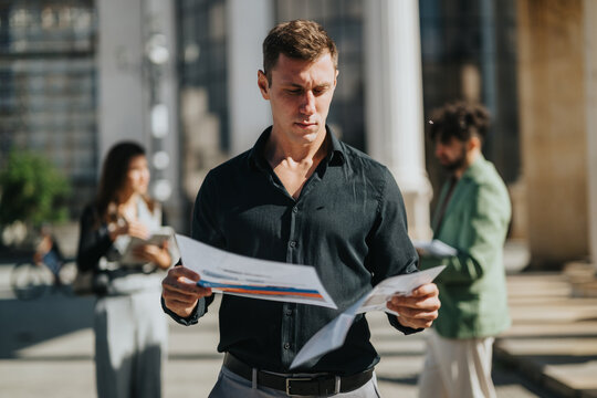 A focused man studies papers outside as two coworkers mingle in the background, conveying a casual, professional vibe amid city life and on-the-go teamwork.