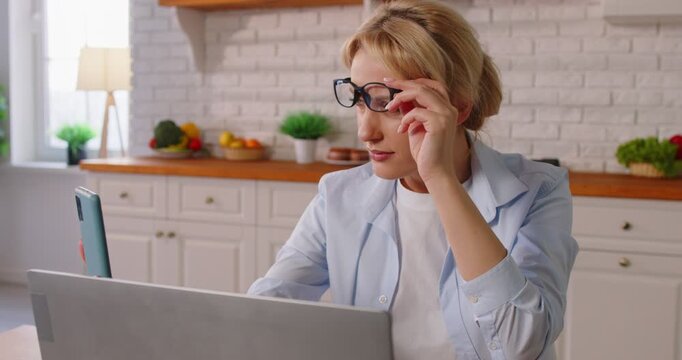 Young woman with farsightedness eye diseases sitting at table in kitchen in eyeglasses trying to read message on smartphone working on laptop at home. Presbyopia and vision problems concept. 4k video