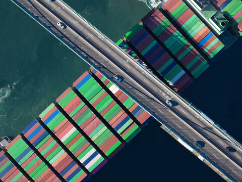 Aerial view of a massive cargo ship, its colorful containers contrasting with the dark blue sea, bisected by a concrete bridge bustling with tiny cars, Los Angeles, California, United States.
