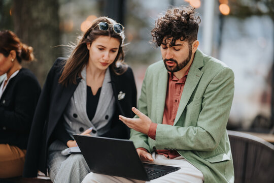 Two well-dressed colleagues sit outdoors, reviewing notes on a laptop. The man gestures as the woman listens, creating a collaborative, modern vibe.