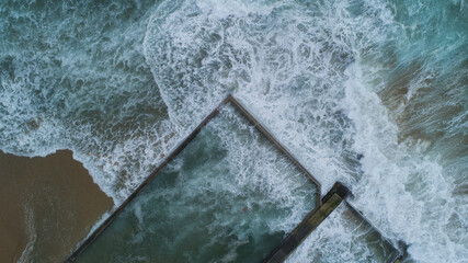 Aerial view of the ocean's forceful waves crashing into the calm, geometric rock pools near the sandy beach, Wollongong, New South Wales, Australia.