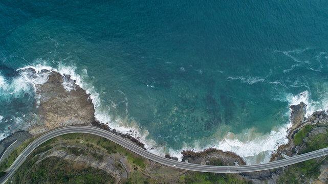 Aerial view of the serpentine Grand Pacific Drive hugging the rugged coastline where the deep blue ocean crashes against the rocks, Wollongong, New South Wales, Australia.
