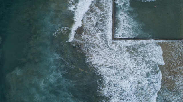 Aerial view of the ocean's tumultuous embrace meets the stark geometry of the North Wollongong Beach pool, a dance of wild surf and human design, Wollongong, New South Wales, Australia.