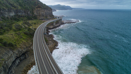 Aerial view of the Sea Cliff Bridge curves along the coast where dark cliffs meet the turquoise sea, Wollongong, New South Wales, Australia.