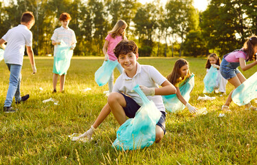 Fototapeta premium Portrait of a happy teenage boy with gloves and trash bag collecting plastic garbage in the summer park with a children volunteers on the background. Environmental pollution and ecology concept.