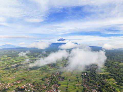 Aerial view of lush rice paddies and verdant trees meet the misty foothills of majestic Mount Merapi under a vast, cloud-strewn sky, Klaten Regency, Central Java, Indonesia.