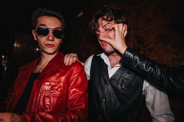 Two men enjoy a nightlife moment, one wearing a red leather jacket and sunglasses, the other in a vest, as a friend covers his face in a playful gesture. Vibrant, candid urban scene.