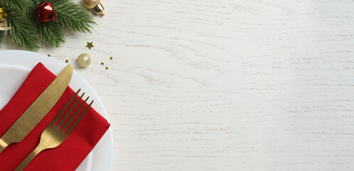 Christmas holiday banner with white plates, gold cutlery, red napkin, evergreen pine tree and ornaments, top view