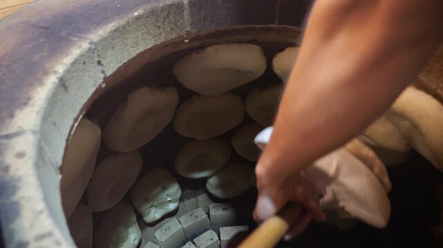 Top view baker's hand glues dough to hot tonir for baking national bread in stone tundyr. Delicious fragrant crispy puri and matnakash. Baking traditional bread.