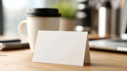 Blank white folded card mockup standing on wooden desk in cozy office with coffee cup and laptop blurred in background.