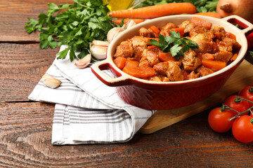 Delicious cooked stew in baking dish and fresh ingredients on wooden table, closeup. Space for text