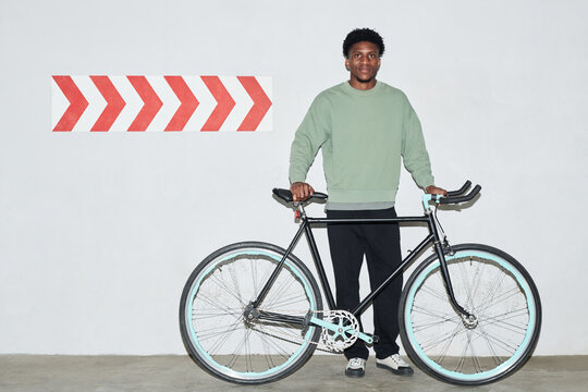 Portrait of young Black man standing indoors holding bicycle, smiling at camera, short curly hair, casual clothing, large red arrow sign on wall in background - Powered by Adobe