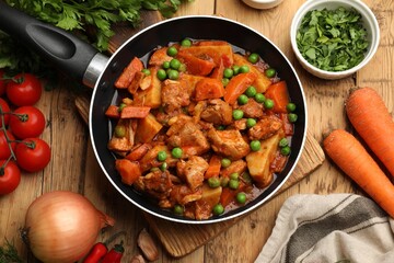Delicious cooked stew in frying pan and ingredients on wooden table, flat lay
