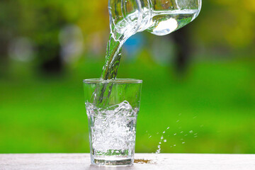 Pouring water into glass from jug at wooden table against blurred green background, closeup