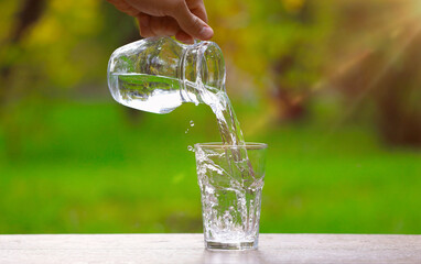 Man pouring water into glass from jug at wooden table against blurred green background, closeup