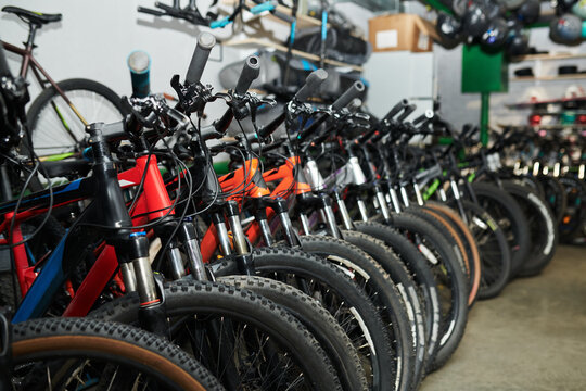 Row of mountain bikes standing in bike shop, handlebars and front wheels aligned closely, various models arranged for sale, equipment and helmets visible in background - Powered by Adobe