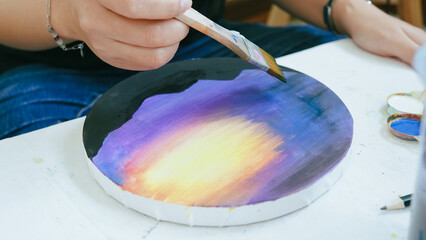 Hand painting a sunset on round canvas using a brush with gradient colors, representing creativity and artistic expression.