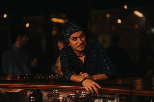 A man wearing a bandana and plaid shirt chats at a dimly lit bar. The scene captures nightlife, social interaction, and a laid-back mood during an evening out with friends. - Powered by Adobe