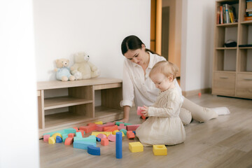 Young happy mother playing game with educational pyramid with cute little child girl in the living room at home. Woman parent spending time with daughter. Family leisure and people concept.