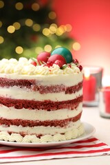Delicious sponge cake with Christmas decor on white table against red background with blurred lights, closeup. Bokeh effect