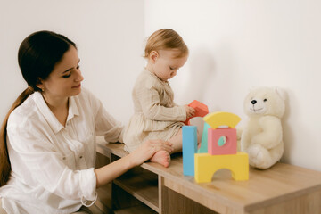 Young happy mother playing game with educational pyramid with cute little child girl in the living room at home. Woman parent spending time with daughter. Family leisure and people concept.